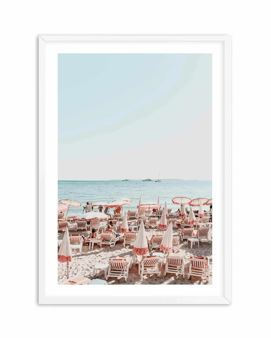 Framed photography art print of a crowded beach scene with white frame and white border. The composition features rows of white and coral pink striped beach chairs and matching umbrellas on light sand, with people relaxing and walking. In the background, the calm turquoise ocean stretches to the horizon with a few yachts and boats visible under a pale blue sky.