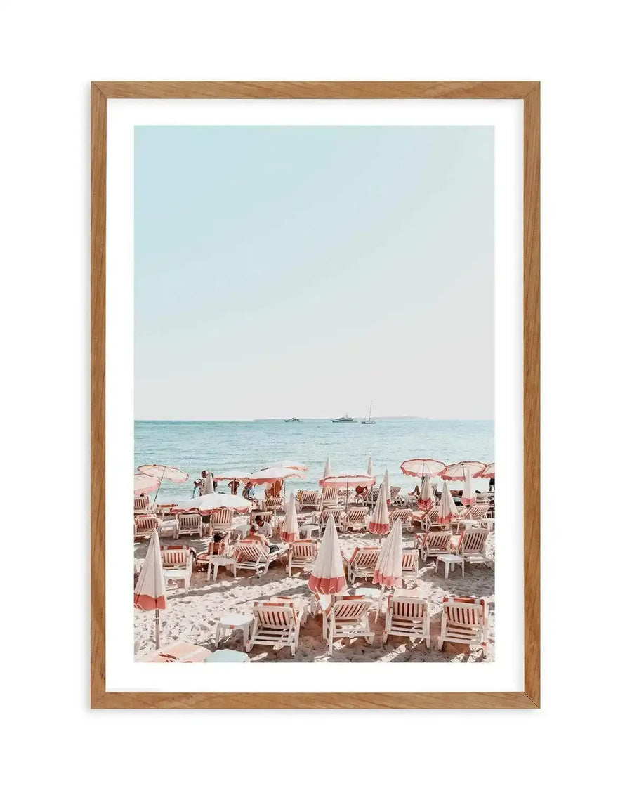 Framed photography art print of a vibrant beach scene with a natural wood frame and white border. The composition features numerous white and pink striped beach chairs and umbrellas on a sandy beach, with people relaxing and swimming in the turquoise ocean. Several boats are visible on the horizon under a clear, pale blue sky.