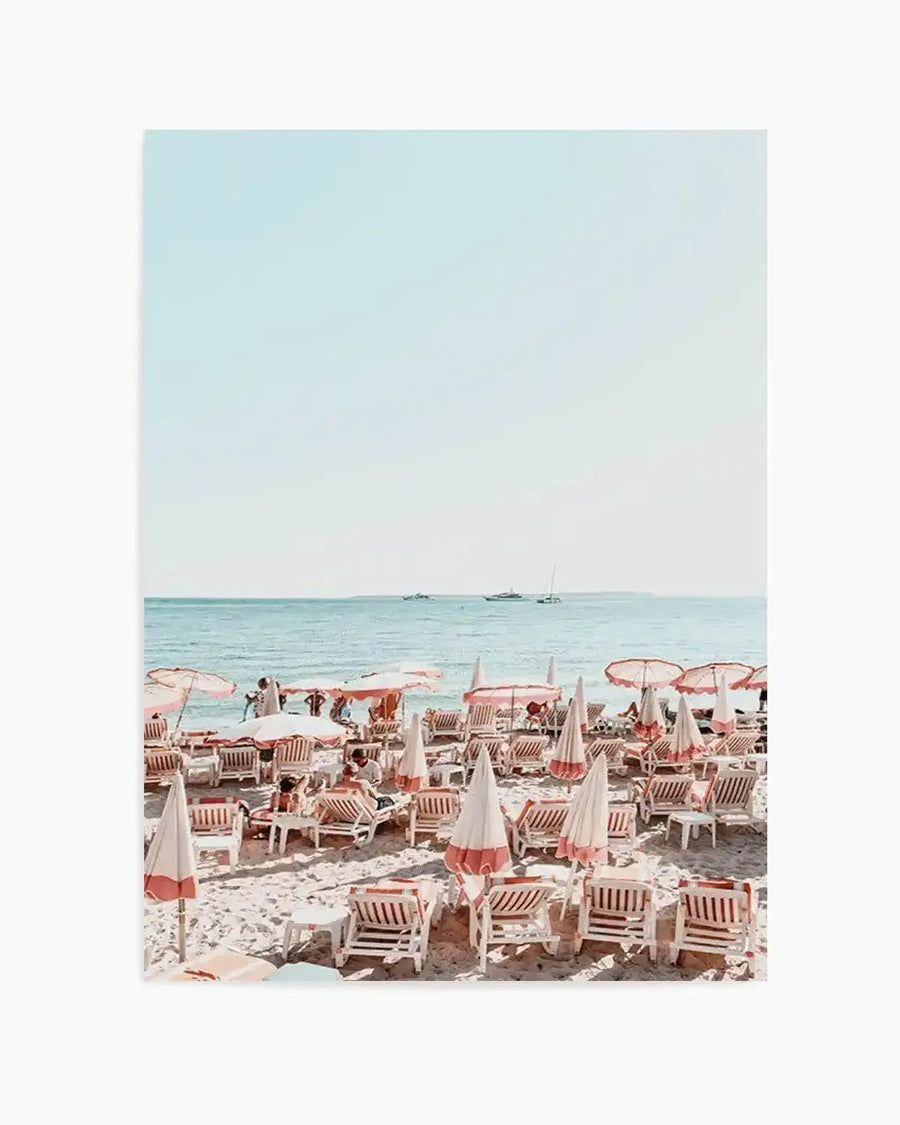 "Photography art print featuring a bustling beach scene with rows of white and coral-pink striped beach umbrellas and lounge chairs on light sand, facing a calm turquoise ocean with distant boats under a pale blue sky."