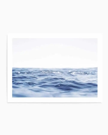 "Photography art print featuring a close-up of ocean water with gentle, rippling waves under a bright, soft white sky. The water is a deep blue with lighter blue highlights and subtle reflections of sunlight on the surface, creating a serene and minimalist composition."