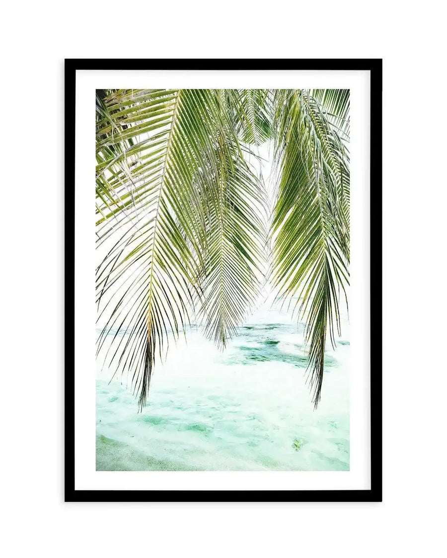 Framed photography art print featuring a tropical beach scene with lush green palm fronds hanging down into the frame from the top, overlooking clear turquoise ocean water with gentle waves. The print has a black frame and a white border.