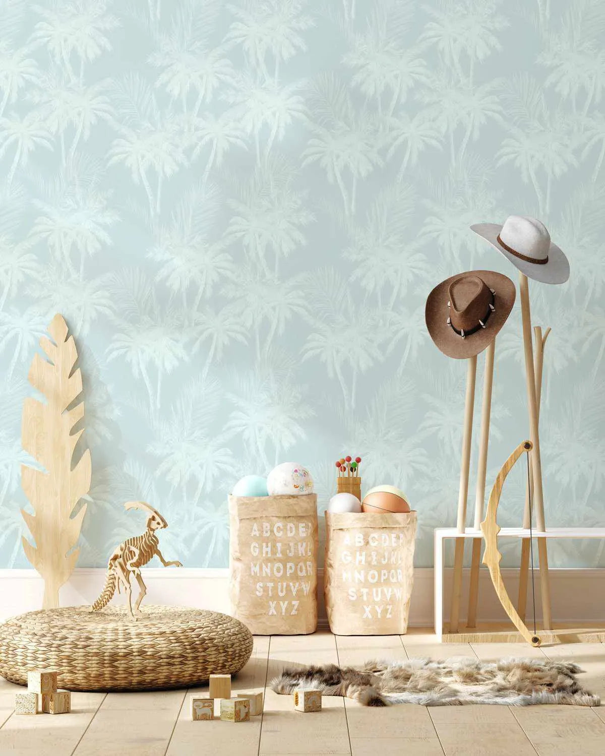 Seaside Palms Wallpaper displayed on a bedroom wall featuring a soft sage green background with repeating palm tree motifs in pale cream. The delicate, hand-painted illustration style shows tall feathered palms creating a tropical pattern. Room styling includes natural wood furniture, woven textiles, straw hats, wooden decorative objects, and neutral coastal accessories arranged on light wood flooring below.