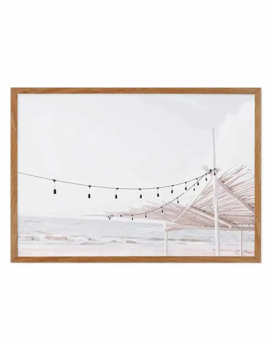 Framed photography art print of a serene beach scene with a string of Edison-style lights hanging over the sand and ocean, displayed in a natural oak frame with white border. The composition features gentle waves on a light grey ocean under a soft white sky, with white wooden structures and palm frond roofs on the right.