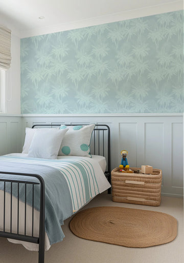 "Seaside Palms wallpaper featuring a repeating pattern of palm trees in a soft, muted sage green against a pale blue background, creating a serene tropical ambiance in a child's bedroom. The wallpaper covers the upper half of the wall, above white wainscoting. A black metal bed frame with white and blue striped bedding, and a woven basket with toys, are visible in the foreground."