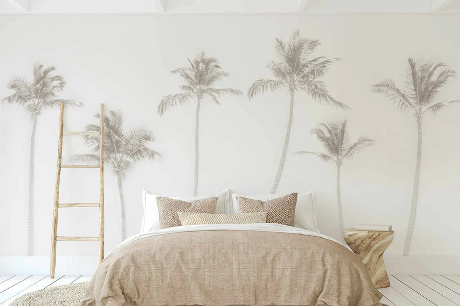 Peel and stick wallpaper featuring a repeating pattern of faded, light grey palm trees on a soft white background, displayed in a minimalist bedroom. The wallpaper covers the wall behind a bed with a textured beige duvet and pillows, and a wooden ladder leans against the wall on the left.