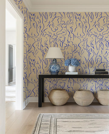 Hallway: Peel and stick wallpaper featuring a repeating pattern of abstract female figures in various poses, rendered in a continuous, flowing royal blue line art style on a warm beige background. The wallpaper is displayed in a hallway with light wood flooring, a black console table with a blue and white patterned lamp, a white vase with blue hydrangeas, and three woven baskets on the floor.