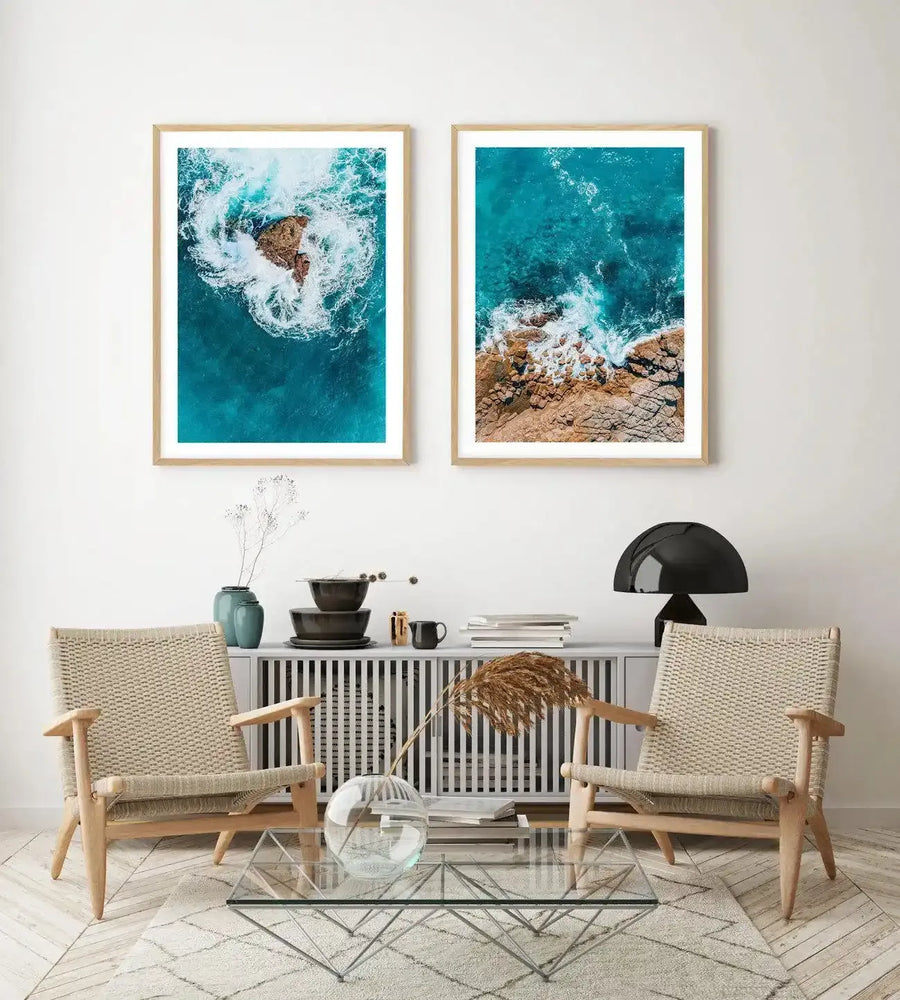 Two framed aerial photography art prints displayed side-by-side above a console table in a minimalist living room. The left print features a single rocky outcrop in turquoise water with white waves crashing around it, viewed from directly above. The right print shows a rugged coastline with brown rocks meeting turquoise water and white waves. Both prints have natural wood frames and white borders. The room includes two woven chairs, a glass coffee table, and a black mushroom lamp.