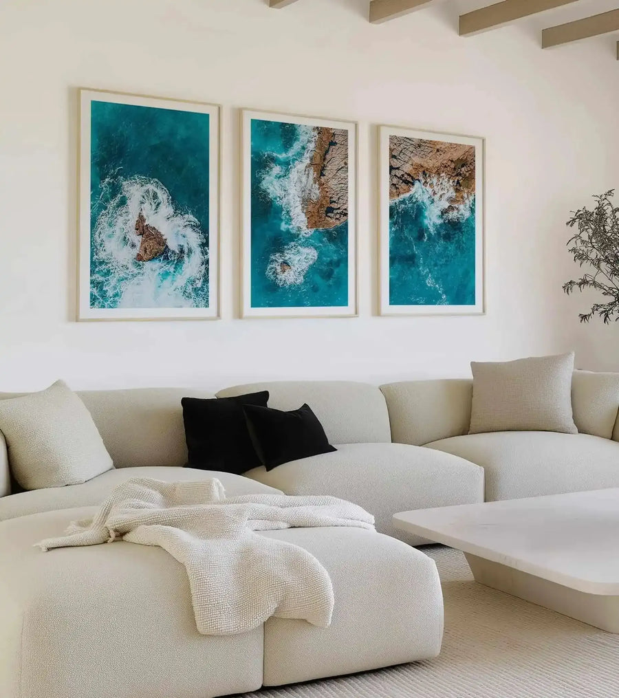 Lifestyle shot of three framed aerial photography art prints displayed above a curved cream sofa in a minimalist living room. Each print depicts a different perspective of turquoise ocean waves crashing against rugged brown rock formations, creating white foamy spray. The prints are framed in natural wood with white borders. The sofa is adorned with cream and black throw pillows and a textured cream throw blanket. A low, rectangular white coffee table sits in front of the sofa.