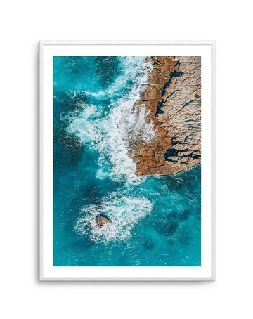Framed aerial photograph of a rugged coastline with turquoise ocean and crashing white waves, displayed in a white frame with white border. The composition captures the vibrant blue and green hues of the water contrasting with the warm brown and tan tones of the rocky shore, with foamy white waves breaking against the land and around a small rock formation in the foreground.