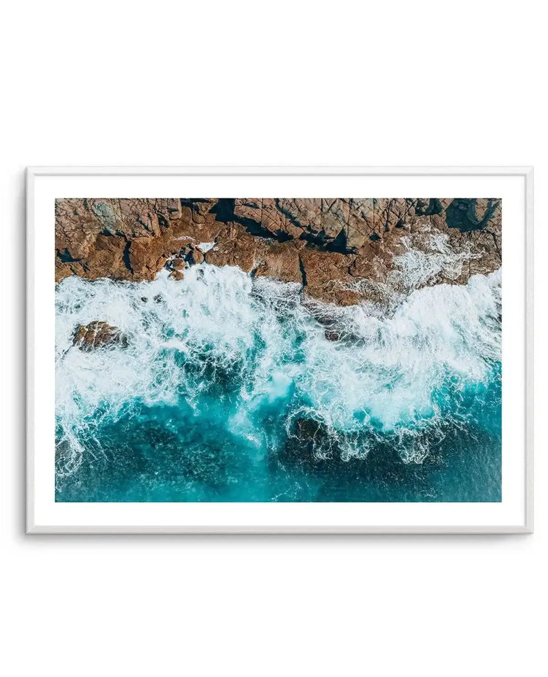 Framed aerial photograph of a rugged coastline with crashing waves and turquoise water, displayed in a white frame with a white border. The composition shows the dark brown and reddish-brown rocks of the headland meeting the vibrant blue and white foam of the ocean, with deeper blue water extending to the bottom of the frame.