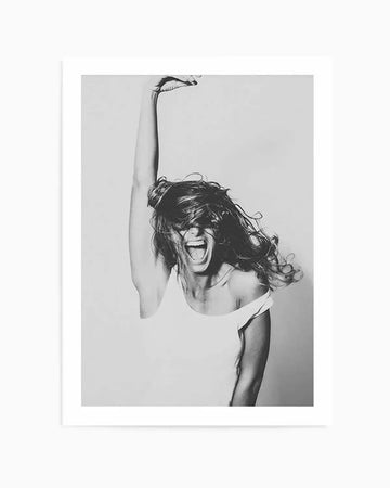 Framed black and white photograph of a woman with her head thrown back, mouth open in a shout or laugh, and hair flying around her face. Her right arm is raised high above her head, and she is wearing a light-colored, off-the-shoulder top. The image captures a moment of energetic joy or excitement, with a natural wood frame and white border.