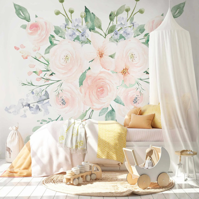 Lifestyle shot of a child's bedroom featuring a peel and stick wallpaper mural with large-scale watercolor flowers in soft pinks, creams, and light blues on a white background. The mural depicts a central bouquet of roses and other blossoms with green leaves and delicate blue accent flowers. The room includes a white bed with yellow and cream bedding, a white sheer canopy, a round woven rug, and wooden toys including a truck and a doll pram.