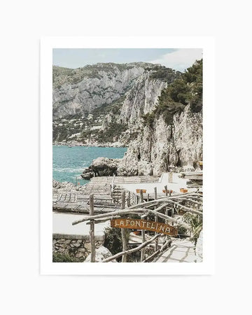 Framed photography art print capturing a scenic coastal view of Capri, Italy, with a natural wood frame and white border. The composition features rugged white cliffs covered in green foliage descending to a turquoise sea, with a small village nestled on the distant hillside. In the foreground, a rustic wooden sign reads "LA FONTELINA" above a path leading to a restaurant by the water.