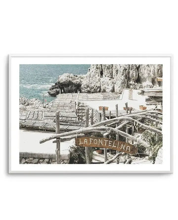 Framed black and white photograph of a coastal restaurant in Capri, Italy, with natural wood frame and white border. The composition features a rustic outdoor dining area with wooden pergolas and a sign reading "LA FONTELINA" in the foreground, with the deep blue sea and rugged white cliffs in the background.