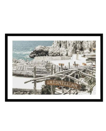 Framed black and white photograph of a coastal restaurant scene with black frame and white border. The composition features a rustic outdoor restaurant with a wooden sign reading "LA FONTELINA" in front of the sea and rocky cliffs. The restaurant has thatched roofs and wooden railings, with potted plants visible.