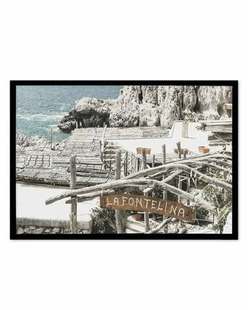 Framed photograph of a coastal scene with a natural wood frame and white border. The composition features a rocky coastline with light blue water, a wooden structure extending over the water, and a sign that reads "LA FONTELINA" in front of a white building with potted plants.