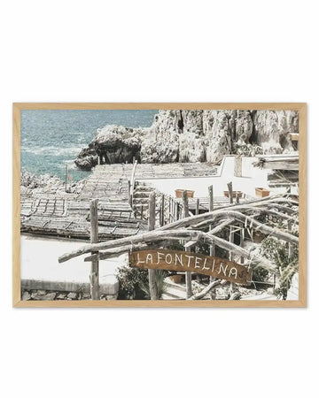 Framed photography art print of a coastal scene in Italy, featuring a white building with a wooden roof and a sign that reads "LA FONTELINA" in front of a rocky cliff and the ocean, displayed in a natural oak frame with white border.