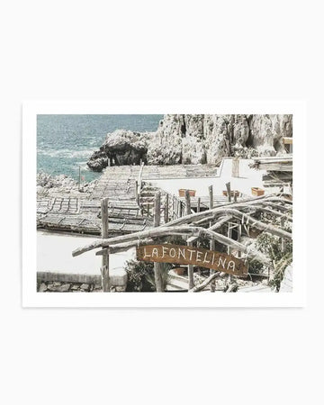 "Photography art print of a coastal scene in Capri, Italy, featuring a restaurant called La Fontelina built into a rocky cliffside overlooking the turquoise sea. The composition captures the rustic charm of the restaurant with its wooden pergolas and white terraces, with a hand-painted wooden sign reading 'LA FONTELINA' in the foreground."