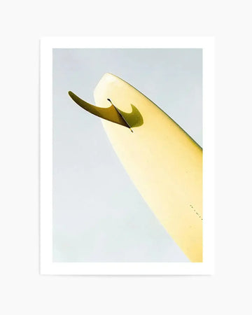 Art print titled "Single Fin Burnt Lemon" by Mario Stefanelli featuring a minimalist illustration of a pale yellow surfboard with a burnt olive-green fin against a soft blue-gray background. The composition is angled diagonally, showing the board's sleek profile and fin detail in sharp focus. The artwork has a clean, graphic quality with soft color gradations. The print is mounted in a white frame with white border.