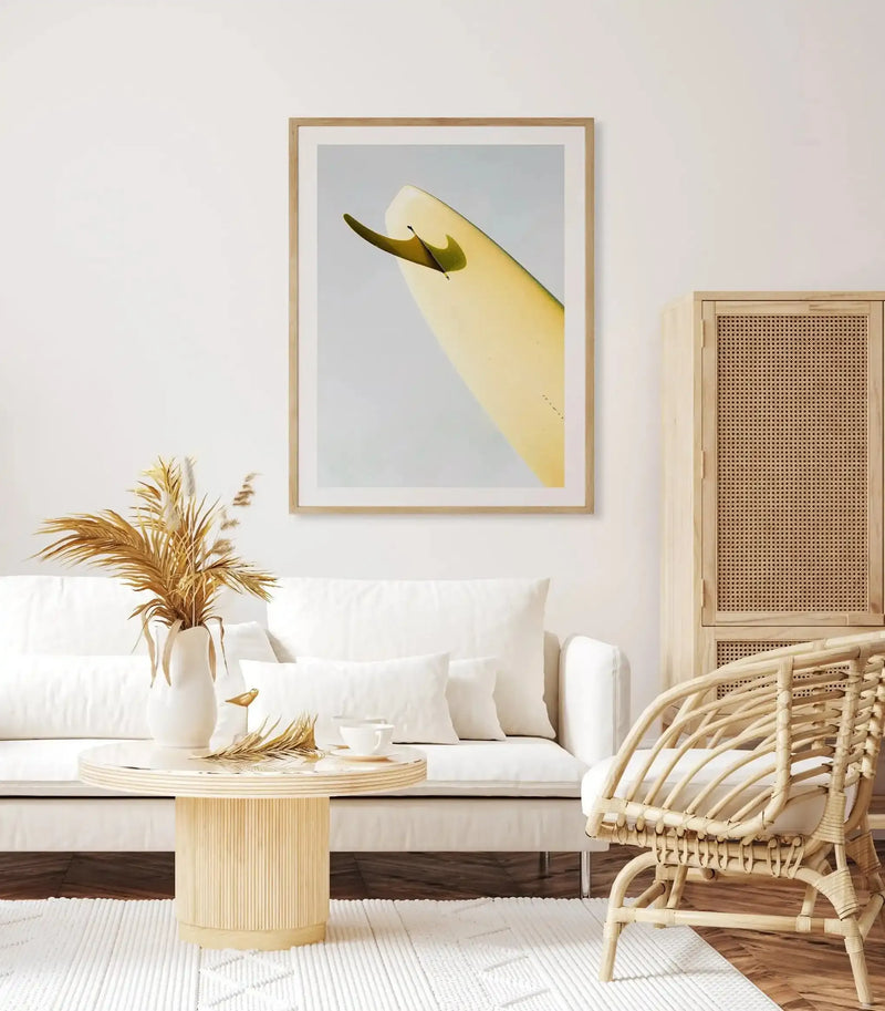 Art print titled "Single Fin Burnt Lemon" by Mario Stefanelli displayed in a natural wood frame with white border on a cream wall above a white upholstered sofa. The artwork features a minimalist illustration of a pale yellow surfboard with a dark olive-green fin against a soft grey-blue background. The print is shown in a styled living room setting with a natural wood side table, dried pampas grass in a white ceramic vase, a woven rattan chair, and a geometric patterned area rug in neutral tones.