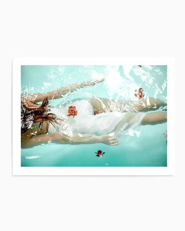 "Art print featuring an overhead photograph of a person floating face-up in clear turquoise water, wearing a white dress. Their long brown hair spreads out around their head, and several red rose petals float on the water's surface around them, creating a serene and ethereal composition."