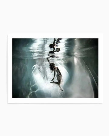 Framed photograph of a person underwater, captured mid-motion with light streaming from above. The person, appearing to be a woman, is depicted in a dark swimsuit, with her body angled upwards and arms extended as if reaching towards the surface. Bubbles surround her, and the water's surface reflects a bright, almost ethereal glow, creating a dynamic and mysterious mood. The print is displayed in a white frame with a white border.