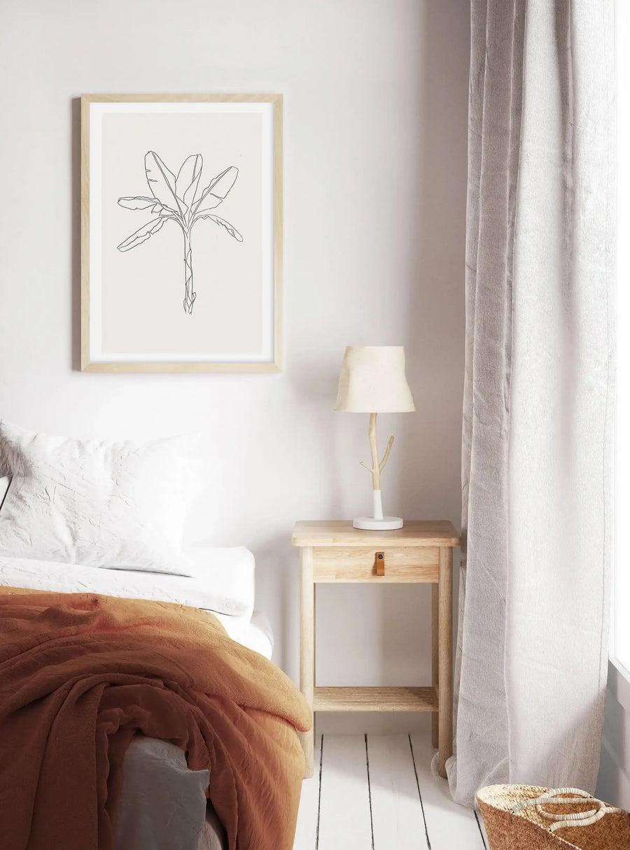 "Framed art print displayed above a bed in a minimalist bedroom. The print features a single line drawing of a palm plant with large leaves and a slender trunk in black on a light cream background, with a natural wood frame and white border. The bed has white bedding and a rust-orange throw, and a light wood bedside table with a lamp and a woven basket on the floor."