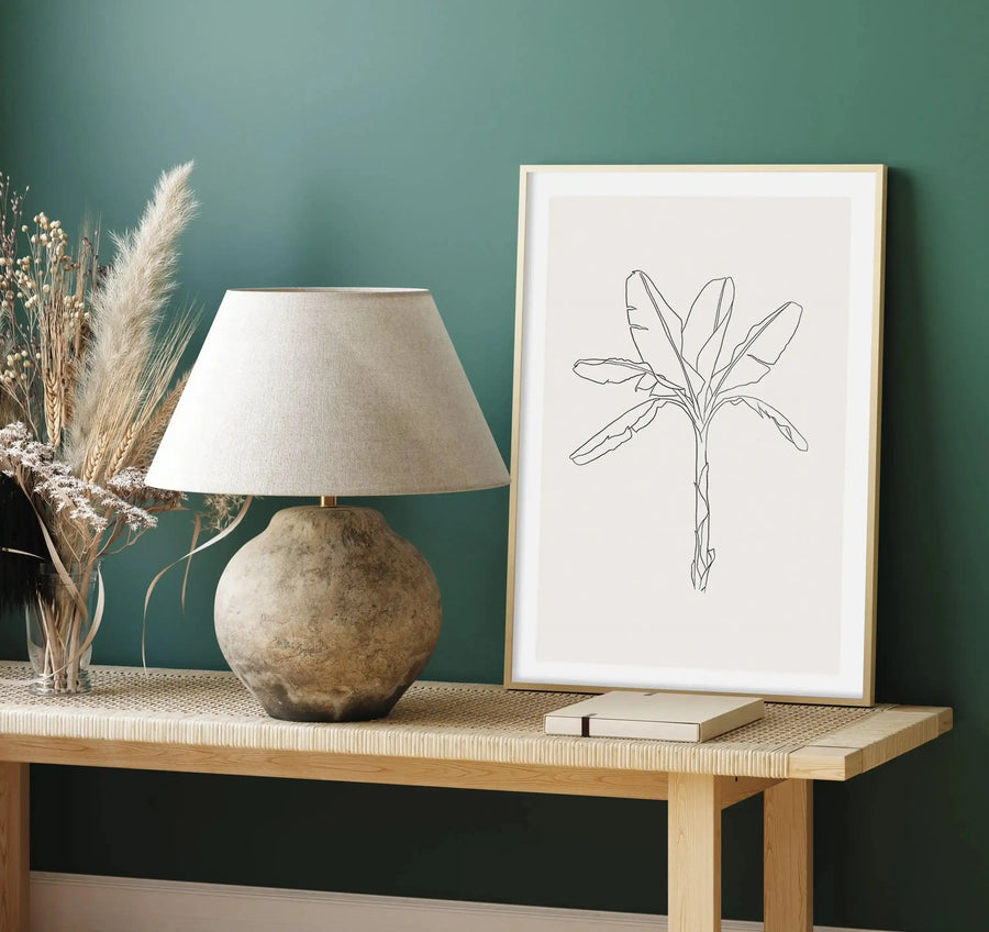 Lifestyle shot of an art print displayed on a wooden console table in a room with dark teal walls. The art print features a minimalist line drawing of a banana palm tree with large leaves and a textured trunk, on a cream background, with a natural wood frame and white border. The console table has a woven top and is adorned with a textured ceramic lamp and a clear vase filled with dried pampas grass and other dried botanicals.