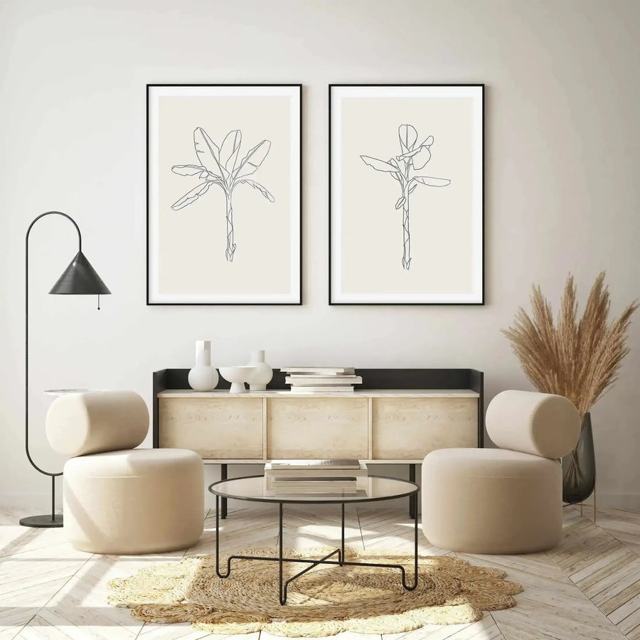Lifestyle shot of two framed art prints displayed above a wooden console table in a minimalist living room. Each print features a single palm tree sketched in black outline on a warm cream background, with a black frame and white border. The room includes two cream upholstered chairs, a black floor lamp, a round glass coffee table on a woven rug, and dried pampas grass in a dark vase.