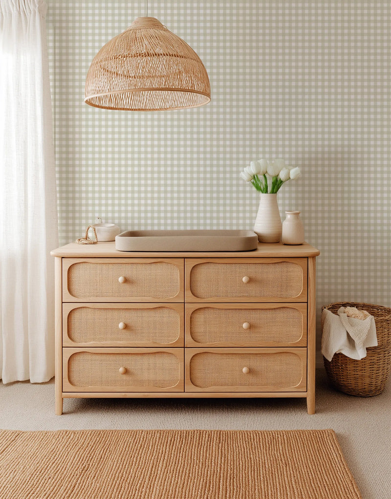 Lifestyle shot of a nursery or bedroom featuring a chest of drawers with a changing pad, a woven basket, and a vase of white tulips against a wall covered in Small Gingham Check New Neutral Green Wallpaper. The wallpaper features a repeating pattern of small, evenly spaced squares in a soft, muted sage green and off-white, creating a classic gingham check design. The room has a light, airy feel with a sheer white curtain and a natural fiber rug.
