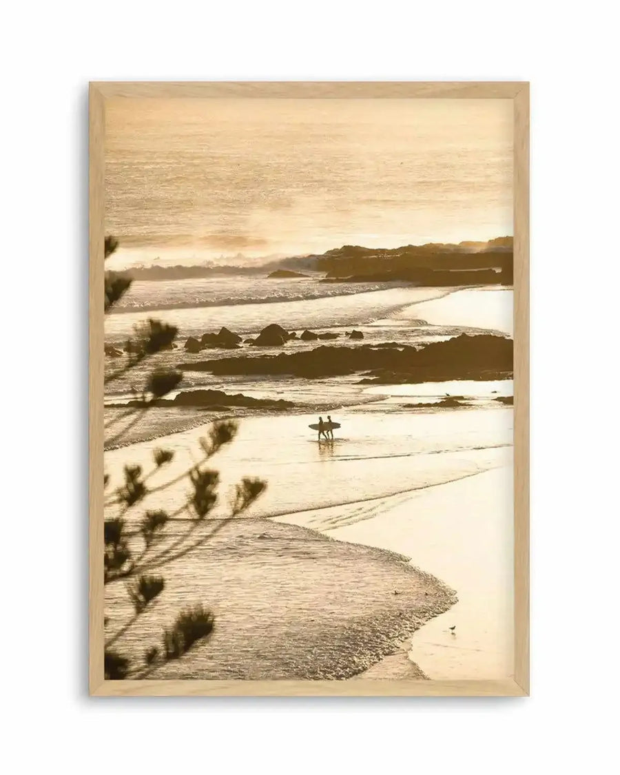 Framed photography art print of a coastal beach scene at sunset with a natural wood frame and white border. The composition features two surfers walking along the wet sand at low tide, with gentle waves breaking on the shore and a warm, golden light illuminating the scene. Silhouetted pine branches are visible in the foreground on the left.