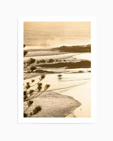 Framed photography art print of a coastal beach scene at sunset, with two surfers walking along wet sand at low tide. The composition features gentle waves and a warm, golden-orange sky stretching to the horizon, with dark rocks scattered along the shoreline. The print is displayed in a white frame with a white border.