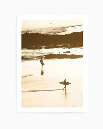 Framed photography art print featuring a golden-hour coastal beach scene with a natural wood frame and white border. The composition captures two silhouetted surfers walking along wet, reflective sand, with gentle waves and rocky outcrops under a warm, glowing sky.