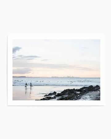 Framed photograph of a coastal beach scene with a white border. The composition features two surfers walking along wet sand at low tide with gentle waves and an overcast sky stretching to the horizon, with a distant city skyline visible.