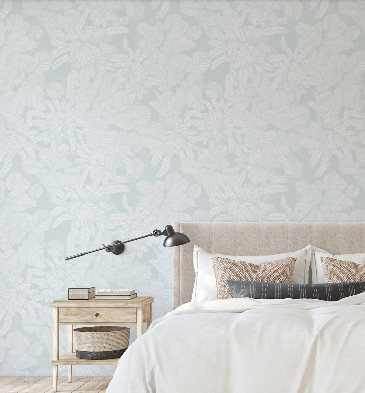 Lifestyle shot of a bedroom featuring Soft Foliage Duck Egg Blue wallpaper behind a bed with a light beige upholstered headboard. The wallpaper has a repeating pattern of various tropical leaves in a soft white outline against a pale duck egg blue background. The bed is dressed with white linen and neutral-toned pillows, and a light wood bedside table with a woven basket sits to the left.