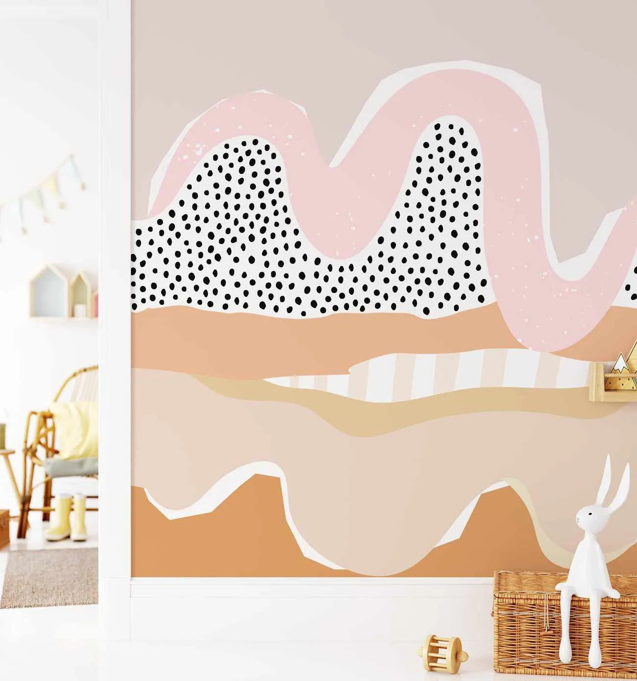 Nursery:
Peel and stick wallpaper featuring an abstract landscape design in a child's room. The mural depicts rolling hills and abstract shapes in soft pink, terracotta, beige, and cream tones, with a large wavy pink shape outlined in white and filled with black polka dots. The wallpaper covers a wall in a nursery with a white rabbit toy sitting on a wicker basket in the foreground.