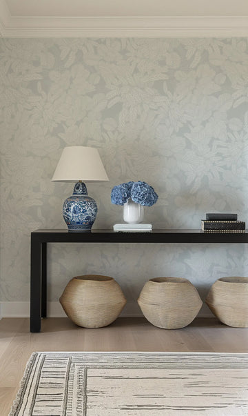 Lifestyle shot of a console table against a wall covered in Soft Foliage | Duck Egg Blue Wallpaper. The wallpaper features a repeating pattern of delicate, hand-drawn tropical leaves in a soft off-white against a muted duck egg blue background. The console table is black and holds a blue and white patterned lamp, a white vase with blue hydrangeas, and a stack of books. Three woven baskets sit on the floor beneath the table, and a striped rug is partially visible in the foreground.