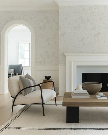 Lifestyle shot of a living room featuring a neutral wallpaper with a soft foliage pattern. The wallpaper has a repeating design of light grey leaves and branches on a slightly darker, warm grey background, creating a subtle, textured look. The room includes a white fireplace, a light-colored armchair with a black metal frame and striped grey pillows, a wooden coffee table with decorative objects, and a cream rug with black striped borders. An arched doorway leads to another room with a window.