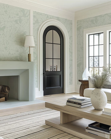 "A living room featuring sage green wallpaper with a soft foliage pattern. The wallpaper has a repeating design of light-coloured leaves with visible vein details, creating a subtle, organic texture. The room includes a light-coloured fireplace, a dark arched door with glass panes, and large windows with black frames. A light wood coffee table with books and a textured white vase with dried foliage sits on a striped rug."