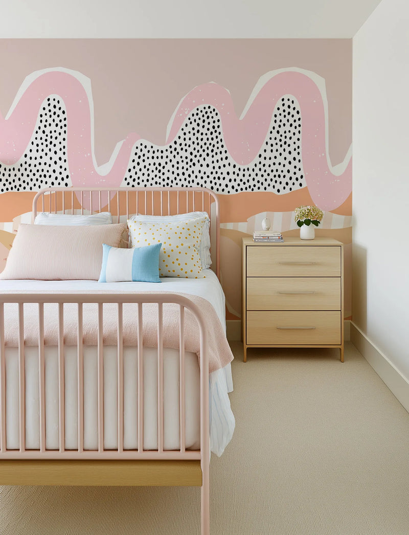 "Soft Outback Mural Wallpaper displayed as a feature wall in a child's bedroom, behind a pink metal bed frame with a light wood base. The wallpaper features an abstract design with large, organic, wavy shapes in soft pink and cream, overlaid with black speckles and white splatters, against a muted peach and light terracotta background. The room includes a light wood bedside table with three drawers, a white vase with flowers, and a light beige textured carpet."