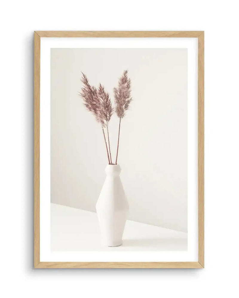 Framed photography art print featuring three stems of dried pampas grass with fluffy, feathery plumes in a soft dusty rose hue, displayed in a white ceramic vase with a faceted design. The vase sits on a white surface against a plain white background, with subtle shadows creating depth. The print is displayed in a natural oak frame with a white border.