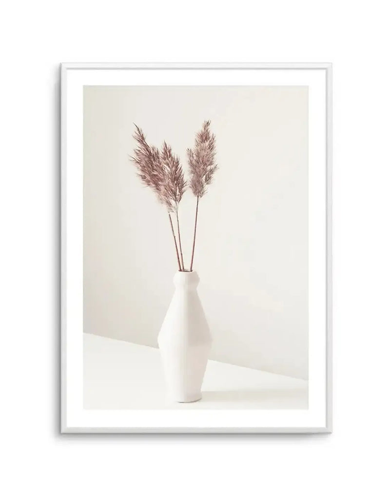 Framed photography art print featuring three stems of dried pampas grass in a white ceramic vase, with a white frame and white border. The pampas grass has feathery, light brown plumes and is arranged centrally in a minimalist composition against a soft, warm white background.