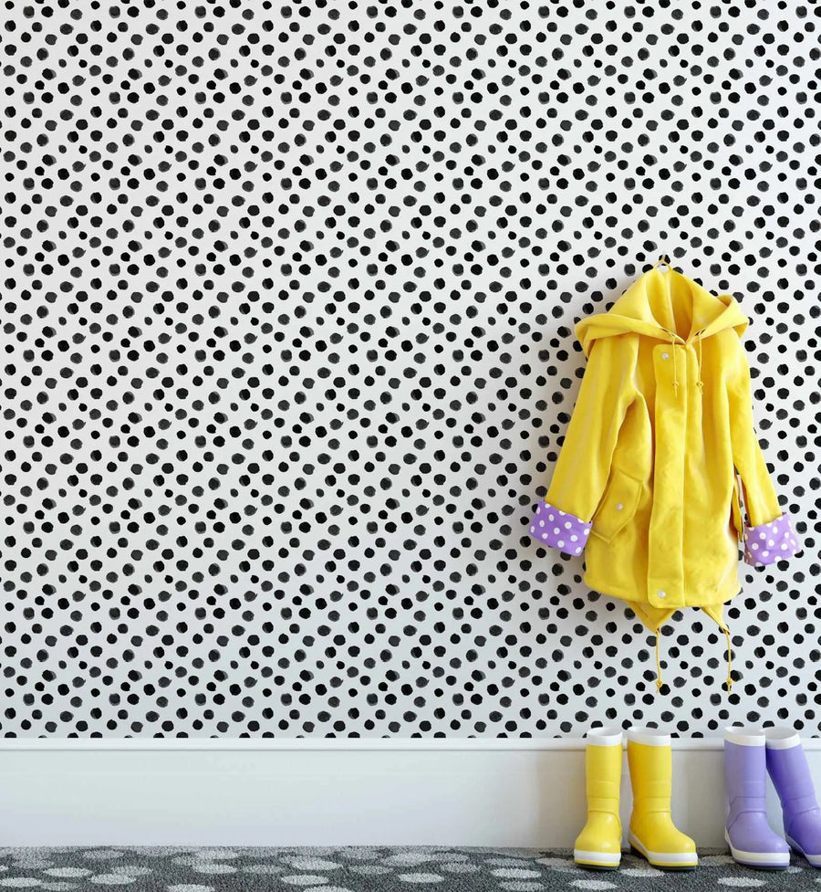 "Peel and stick wallpaper featuring a repeating pattern of hand-painted black dots of varying sizes on a white background. The dots have a loose, organic watercolour quality with visible brushstroke texture. A yellow raincoat with purple and white polka dot cuffs hangs on the wall, and two pairs of rain boots, one yellow and one lavender, sit on a grey patterned rug below."