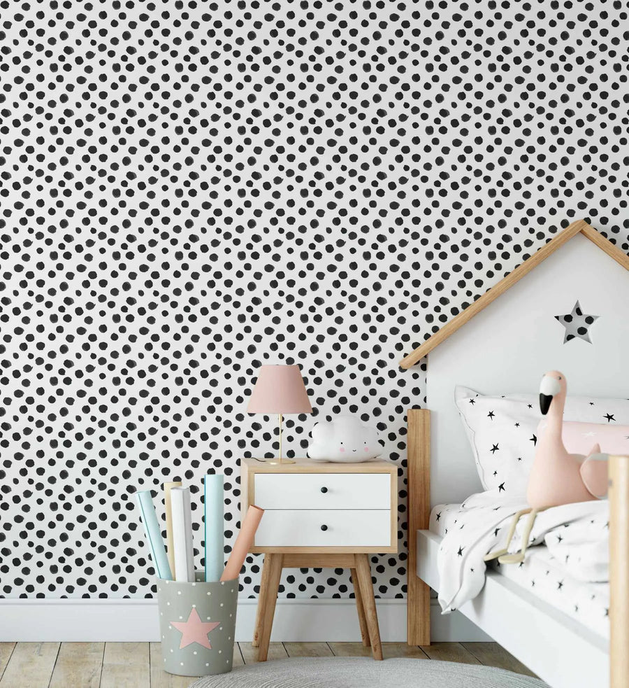 "A child's bedroom featuring a wall covered in black and white polka dot wallpaper. The wallpaper has an organic, hand-painted quality with irregular black dots on a crisp white background. A light wood and white house-shaped bed frame is visible on the right, with a white bedside table and a pink lamp. A grey bucket with a pink star and rolled papers sits on the floor."