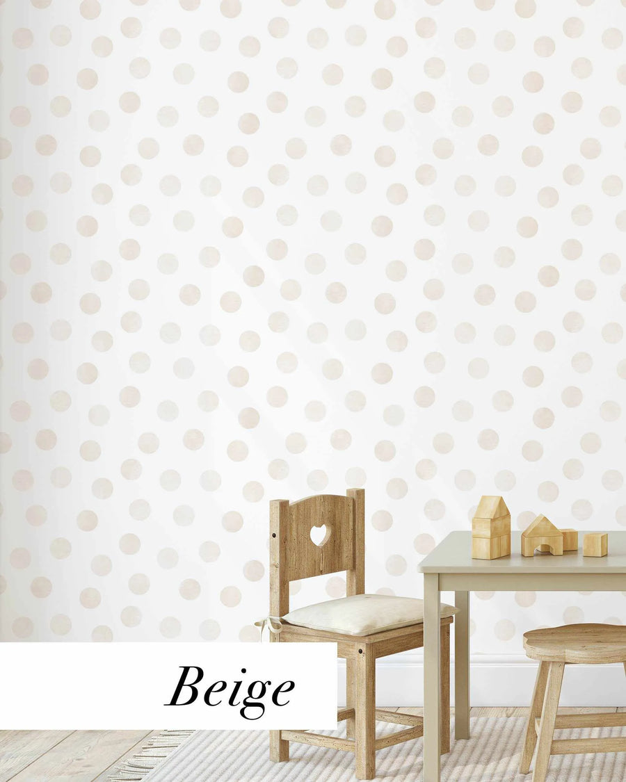 "Peel and stick wallpaper featuring a repeating pattern of hand-painted polka dots in a soft, muted beige on a bright white background. The dots have a slightly irregular, watercolour-like texture and are evenly spaced across the surface. A small wooden chair with a heart cut-out and a light grey table with wooden building blocks are visible in the foreground."