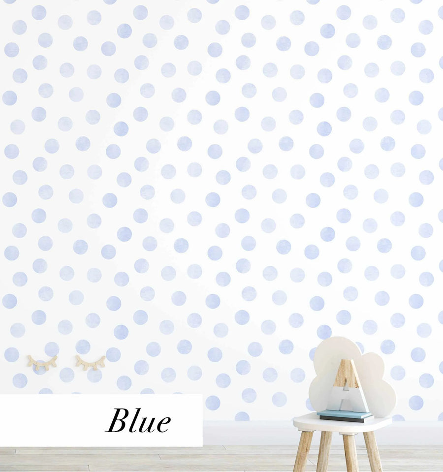 "Peel and stick wallpaper featuring a repeating pattern of hand-painted polka dots in a soft, muted periwinkle blue on a clean white background. The dots have a subtle watercolor texture and are arranged in a slightly irregular, scattered grid. The bottom left corner shows two wooden decorative eyelashes, and the bottom right shows a small white cloud-shaped stool with a wooden letter 'A' and two books."
