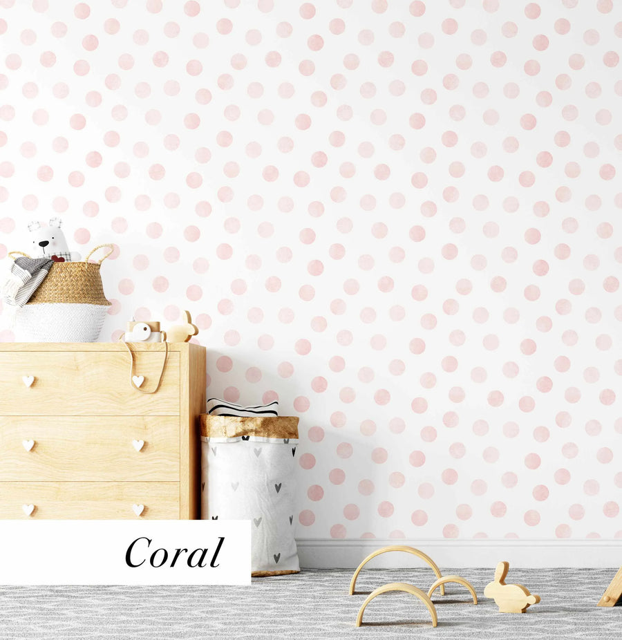 Lifestyle shot of a child's room featuring peel and stick wallpaper with a repeating pattern of hand-painted polka dots in a soft coral pink on a white background. The wallpaper has a watercolour texture with visible brushstrokes. A light wood dresser with heart-shaped knobs and a woven basket with a teddy bear are visible on the left, while a white baseboard and a grey patterned rug are at the bottom. Wooden toys are scattered on the rug.