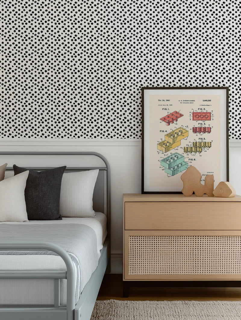 Lifestyle shot of a bedroom featuring Spots & Dots Small Wallpaper on the wall behind a bed with a grey metal frame. The wallpaper has a repeating pattern of irregular, hand-painted black dots on a white background, creating a playful, organic feel. A framed art print of a LEGO patent diagram hangs above a light wood bedside table with a woven cane drawer front.
