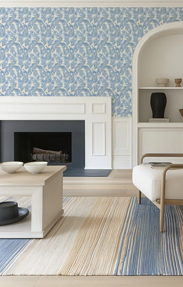 Lifestyle shot of a living room featuring a wall covered in Spotted Palms Wallpaper. The wallpaper has a repeating pattern of hand-painted palm fronds in varying shades of light blue and indigo on a soft cream background, creating a fresh, tropical feel. The wall includes a white fireplace mantel and built-in shelving with decorative items. A light wood coffee table with white bowls and a striped rug in blue, cream, and beige are visible in the foreground.