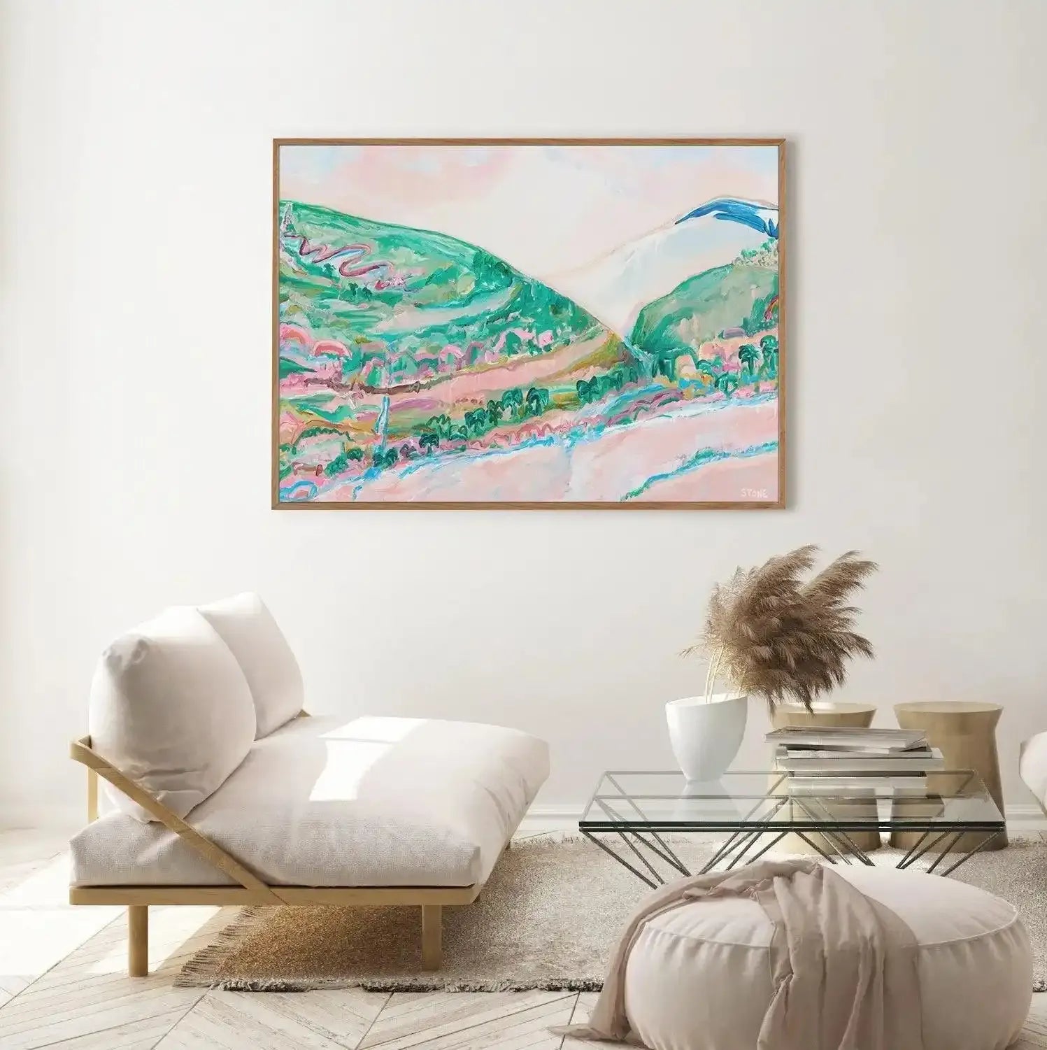Large framed canvas art print titled "Springtime in the Yarra" displayed above a minimalist white chaise lounge in a bright living room. The painting features a vibrant, impressionistic landscape with rolling green hills, a winding pink road, and a pale pink sky. Lush green trees and a blue river are visible at the base of the hills. The artwork is framed in natural wood. The room includes a glass coffee table with dried pampas grass in a white vase, and a round white ottoman with a draped beige throw.